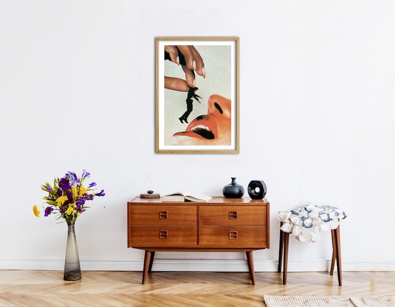 May include: A framed surrealist art print featuring a large hand holding a tiny person above a giant open mouth. The artwork uses a collage style with realistic skin tones, a black silhouette figure, and a light background, all encased in a wooden frame.