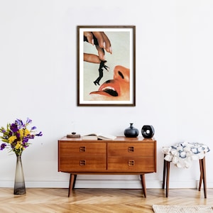May include: A framed surrealist art print featuring a large hand holding a tiny person in a black suit above a human face with an open mouth. The artwork uses a collage style with warm skin tones, black clothing, and a white background.
