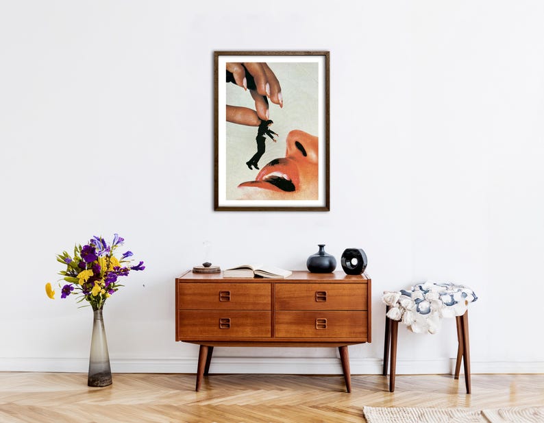 May include: A framed surrealist art print featuring a large hand holding a tiny person in a black suit above a human face with an open mouth. The artwork uses a collage style with warm skin tones, black clothing, and a white background.