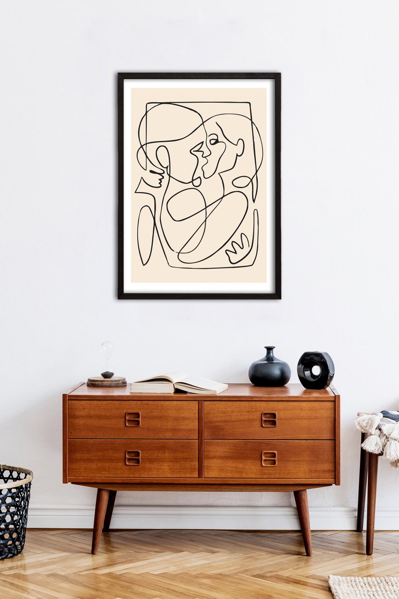 May include: A black and white line drawing of two people embracing, framed in a black frame and hanging on a white wall above a wooden dresser.