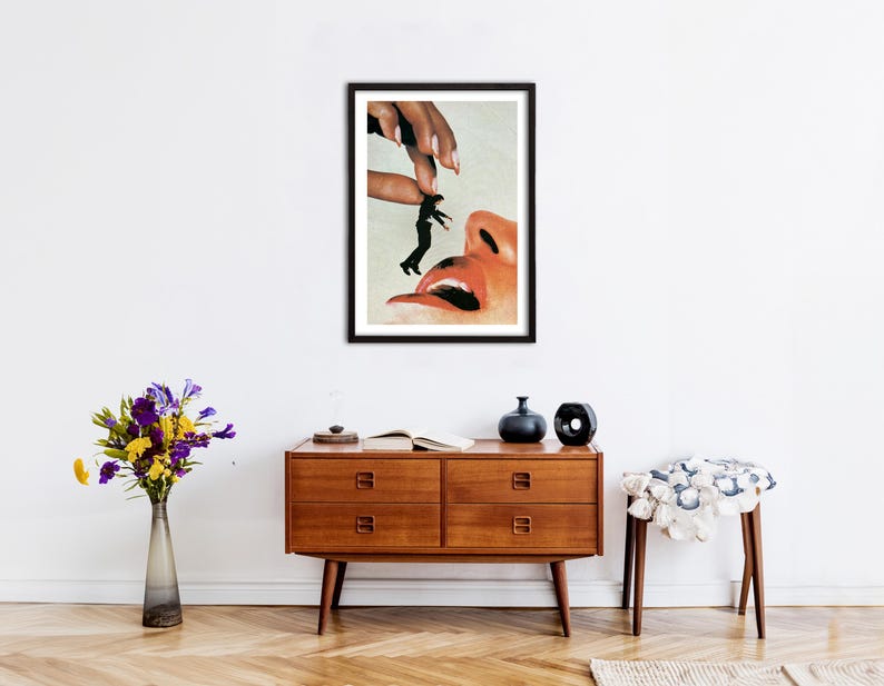 May include: A framed surrealist art print featuring a large hand holding a tiny person in a black suit above a human face with an open mouth and visible tongue. The artwork uses a collage style with warm skin tones and high contrast black elements.