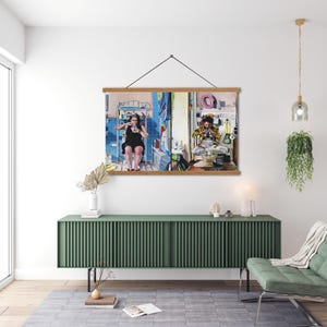 May include: A framed print of a split-screen image showing two people in separate rooms. The person on the left is wearing a black dress and is sitting on a toilet. The person on the right is wearing a yellow and black patterned shirt and is sitting on a toilet. The print is hanging on a green wall above a green cabinet.
