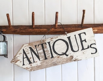 May include: A weathered wooden sign with the word "ANTIQUE" painted in black letters hangs from a rustic wooden rack with hooks. A small lantern is hanging on the left side of the rack.