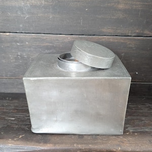 Könnte beinhalten: Eine silberne Metalldose mit Deckel, der Deckel ist leicht geöffnet. Die Dose steht auf einer dunklen Holzoberfläche.