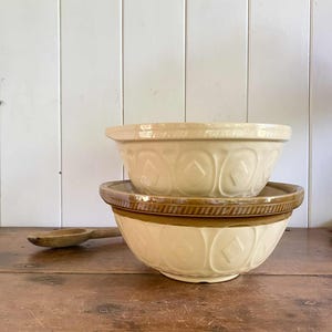 Puede incluir: Dos cuencos de cerámica color crema con un patrón de diamantes, uno apilado encima del otro. El cuenco superior es más pequeño. Una cuchara de madera descansa sobre la superficie de madera junto a los cuencos. Los cuencos tienen un borde marrón.