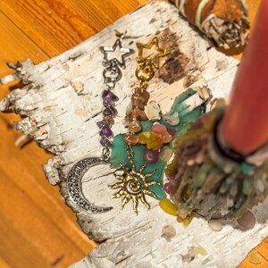 May include: Two charm necklaces with celestial designs. One features a silver crescent moon and amethyst beads, the other a gold sun pendant and gemstone beads. The charms hang on a piece of birch bark.