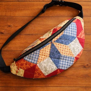 May include: A patchwork fanny pack with a black zip and adjustable strap. The bag features a variety of fabric patterns in red, blue, yellow, and white. The bag is set against a wooden background.
