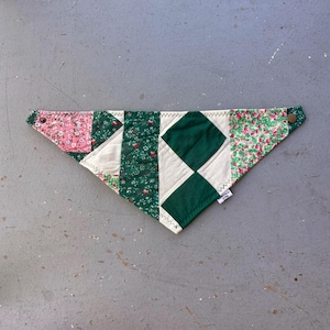 May include: A patchwork bandana featuring floral and geometric patterns in pink, green, and white. The triangular bandana has snap closures at each end. The fabric is stitched together with a zig-zag stitch.