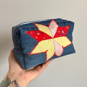 Peut inclure: Une pochette zippée en tissu denim avec un motif patchwork en forme d'étoile. L'étoile présente des accents de tissu rouge, jaune et à motifs. La pochette est tenue dans une main.