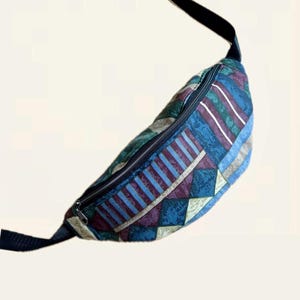May include: A vintage fanny pack featuring a geometric pattern in blue, purple, green, and beige. The bag has a black zip and strap. The design includes stripes, triangles, and other shapes.