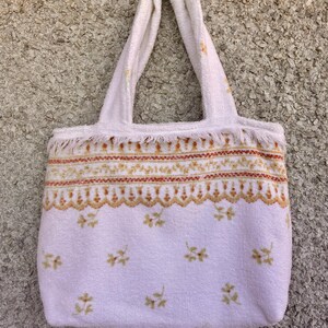 May include: A white tote bag made from a soft, textured material. The bag features a decorative band with orange and yellow patterns, and small floral accents. The handles are made of the same material.