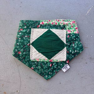 May include: A quilted, pentagon-shaped item with a green floral pattern and a central green diamond. The item has a white square with a green diamond in the centre. A small tag with the text "SLOW POKED GOODS" is attached.
