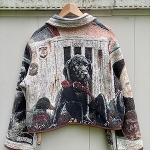 May include: A woven tapestry jacket featuring a prominent black Labrador retriever wearing a red collar. The design includes a barn-like background, various circular emblems, and the text CROWE printed near the bottom hem. The jacket has a collar.