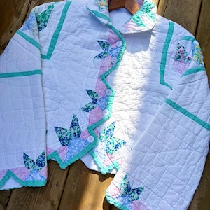 May include: A white quilted jacket with floral and geometric patterns. The jacket has a scalloped edge with pink and blue floral accents, and is trimmed with mint green. It has a button-up front and a collared neckline.