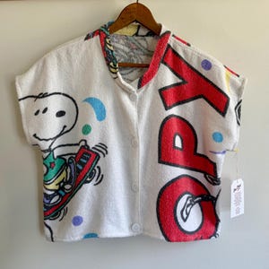 May include: White button-up shirt featuring a Snoopy graphic and the word "SPORT" in red. The shirt has a colourful collar and is decorated with blue, green, and purple polka dots. The shirt is hanging on a wooden hanger.