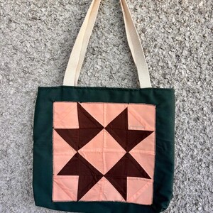 May include: A green tote bag with cream-coloured handles. The bag features a central square design with a patchwork pattern of brown and peach-coloured triangles, creating a star-like motif. The bag is made of fabric.