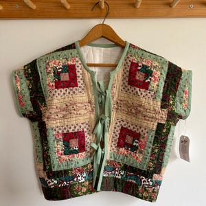 Peut inclure: Un gilet matelassé avec un motif patchwork. Le gilet présente des carrés de tissu rouge, vert et à motifs floraux, avec une bordure et des liens vert clair. Il est suspendu à un cintre en bois contre un mur blanc.