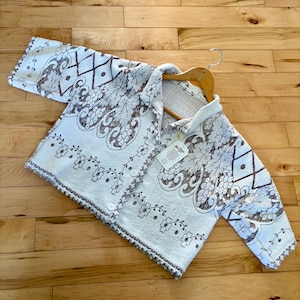 May include: A white button-down shirt with a floral and geometric pattern in brown and beige. The shirt is hanging on a wooden hanger. The shirt has a scalloped edge and a tag.