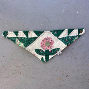Peut inclure: Bandana pour animaux de compagnie matelassé fait à la main avec un motif floral. Le bandana présente une fleur centrale avec des pétales roses et un centre vert, entourée de feuilles vertes et d'un fond blanc. Les bords sont garnis de triangles verts et blancs.