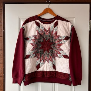 Peut inclure: Un sweat-shirt bordeaux avec un grand motif d'étoile en patchwork blanc et rouge sur le devant. L'étoile est composée de différents motifs floraux et est entourée d'un fond blanc.