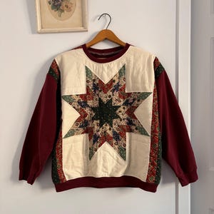 May include: A burgundy sweatshirt featuring a quilted star design. The star is crafted from patterned fabrics in green, red, and blue hues. The sleeves are solid burgundy, and the neckline is trimmed in burgundy. The sweatshirt is hanging on a wooden hanger.