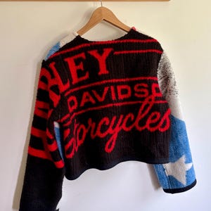 May include: A black, red, and blue fleece jacket featuring the text Harley Davidson Motorcycles in red lettering. The jacket has a textured, fuzzy material with a star pattern on the sleeves and a cropped hemline, hanging on a wooden hanger.