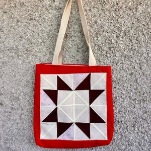 Peut inclure: Sac fourre-tout rouge avec des anses crème. Le sac présente un motif carré matelassé avec un motif d'étoile en blanc, bordeaux et rose clair. Le sac est en tissu et a un design simple et fonctionnel.