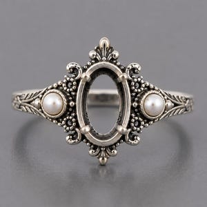 May include: A silver ring featuring an empty central oval setting surrounded by intricate metal scrollwork and two small, round white pearls set on either side of the main frame. The band has a textured, leaf-like pattern along the sides.