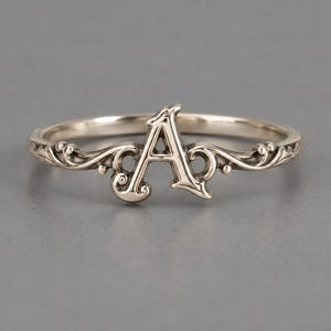 Könnte beinhalten: Ein Ring aus silberfarbenem Metall mit einem zentralen Großbuchstaben A. Das Band ist mit komplizierten, erhabenen Schnörkeln und wirbelnden Filigranmustern verziert, die sich vom Buchstaben aus entlang der Ringseiten erstrecken und ein durchgehendes Design bilden.