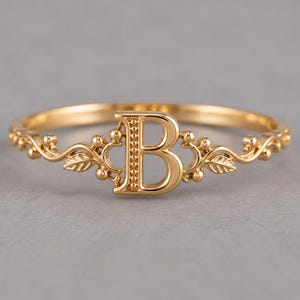 以下が含まれることがあります： 中央に大文字の「B」をあしらったゴールドトーンのリング。リングのバンド部分は、文字の両サイドに小さな葉や球状の装飾が施された左右対称のフィリグリーデザインで、細く磨き上げられた金属製のバンドが特徴です。