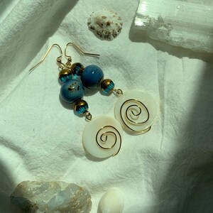 May include: Handmade earrings featuring a combination of blue and white beads with gold accents. The earrings have a unique design with a spiral detail on the white shell-shaped pendants. The jewelry is displayed on a white fabric background.