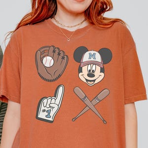 May include: A rust-coloured t-shirt with a graphic print of Mickey Mouse wearing a baseball cap, a baseball glove, a baseball bat, and a foam finger with the number 1 on it.