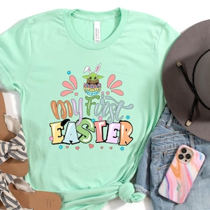 Puede incluir: Una camiseta verde claro con el texto "my first EASTER" en letras coloridas. La camiseta presenta un gr&aacute;fico de un personaje con orejas de conejo. La imagen tambi&eacute;n incluye sandalias, un sombrero y pantalones cortos de mezclilla.