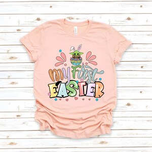 Puede incluir: Una camiseta rosa claro con las palabras "My First Easter" en letras coloridas y juguetonas. El dise&ntilde;o incluye un personaje de dibujos animados en un huevo de Pascua con orejas de conejo. La camiseta es de material suave.