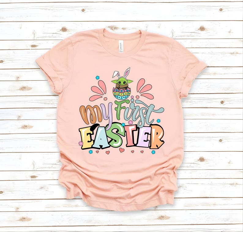 Puede incluir: Una camiseta rosa claro con las palabras "My First Easter" en letras coloridas y juguetonas. El dise&ntilde;o incluye un personaje de dibujos animados en un huevo de Pascua con orejas de conejo. La camiseta es de material suave.