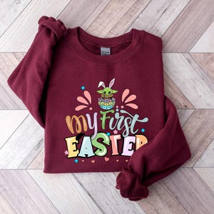 Puede incluir: Una sudadera granate con el texto "my first EASTER" en letras coloridas. El dise&ntilde;o incluye un personaje de dibujos animados con orejas de conejo y un huevo de Pascua. La sudadera es de un material suave.