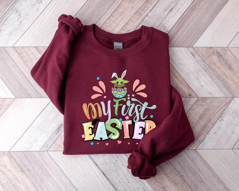 Puede incluir: Una sudadera granate con el texto "my first EASTER" en letras coloridas. El dise&ntilde;o incluye un personaje de dibujos animados con orejas de conejo y un huevo de Pascua. La sudadera es de un material suave.