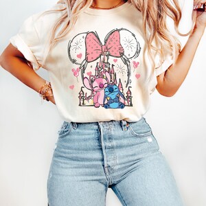 May include: Cream-coloured t-shirt with a pink bow and Mickey Mouse ears design. The shirt features a graphic of a castle, pink hearts, and cartoon characters. The shirt is paired with light blue jeans.
