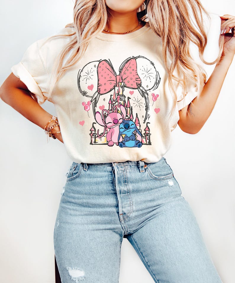 May include: Cream-coloured t-shirt with a pink bow and Mickey Mouse ears design. The shirt features a graphic of a castle, pink hearts, and cartoon characters. The shirt is paired with light blue jeans.