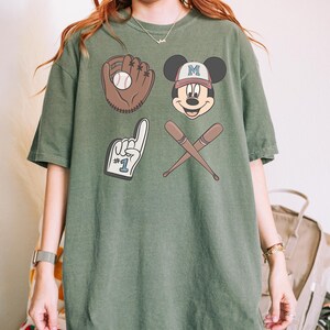 May include: A green t-shirt with a baseball theme featuring Mickey Mouse wearing a baseball cap with the letter "M" on it, a baseball glove with a ball, a baseball bat and a foam finger with the number "1" on it.