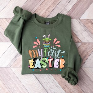 Puede incluir: Una sudadera verde oliva con el texto "my first Easter" en letras coloridas. El gr&aacute;fico incluye un personaje de dibujos animados con orejas de conejo en un huevo de Pascua. La sudadera tiene mangas largas y cuello redondo.
