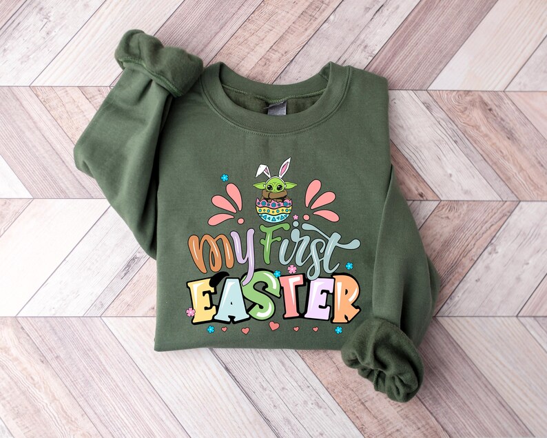 Puede incluir: Una sudadera verde oliva con el texto "my first Easter" en letras coloridas. El gr&aacute;fico incluye un personaje de dibujos animados con orejas de conejo en un huevo de Pascua. La sudadera tiene mangas largas y cuello redondo.