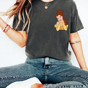 May include: Dark gray t-shirt featuring a cartoon image of Belle from Beauty and the Beast. Belle is depicted with brown hair and a yellow dress. The shirt is paired with blue jeans and white sneakers, creating a casual, everyday look.
