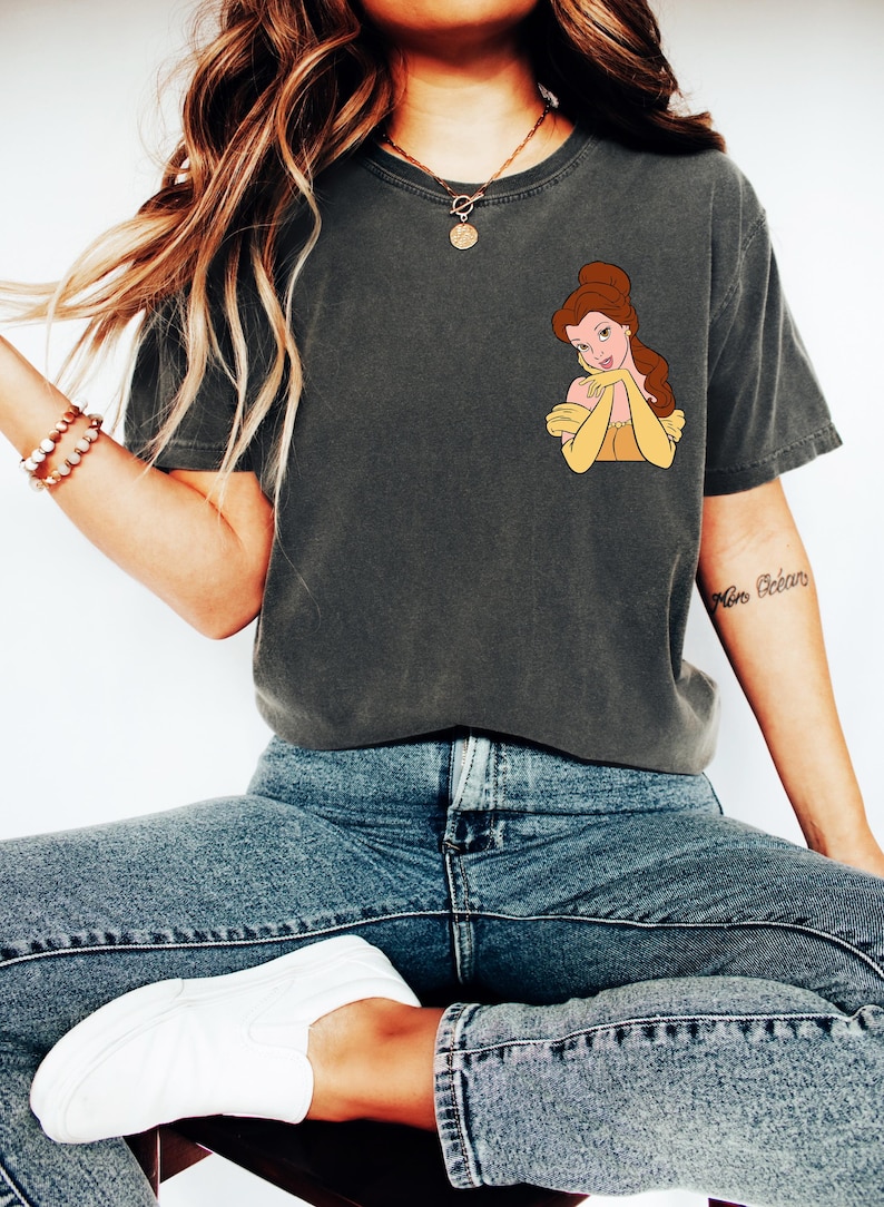 May include: Dark gray t-shirt featuring a cartoon image of Belle from Beauty and the Beast. Belle is depicted with brown hair and a yellow dress. The shirt is paired with blue jeans and white sneakers, creating a casual, everyday look.