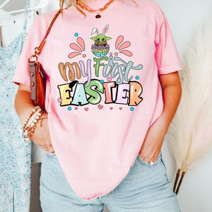 Puede incluir: Camiseta rosa con la frase "My First Easter" en letras coloridas. El dise&ntilde;o incluye un personaje de dibujos animados Grogu en un huevo de Pascua con orejas de conejo. La camiseta es de estilo casual y de manga corta.