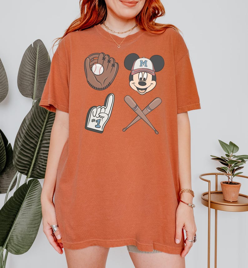 May include: A rust-coloured t-shirt with a graphic print of Mickey Mouse wearing a baseball cap, a baseball glove, a baseball bat, and a foam finger with the number 1 on it.