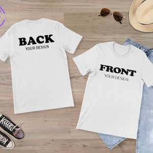 May include: Two white t-shirts with black text that reads "BACK YOUR DESIGN" and "FRONT YOUR DESIGN". The shirts are displayed on a wooden surface with a pair of blue jeans, sunglasses, a hat, and a pair of black sneakers.