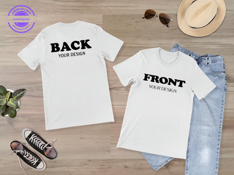 a pair of white t-shirts with the text "BACK YOUR DESIGN" printed on them, along with a pair of jeans and a hat. There are also sunglasses and a pair of sneakers placed on the wooden surface.