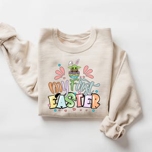 Puede incluir: Sudadera beige con el texto "my first EASTER" en letras de colores. El gr&aacute;fico presenta un personaje de dibujos animados con orejas de conejo en un huevo de Pascua. Acentos rosas y azules rodean el texto.