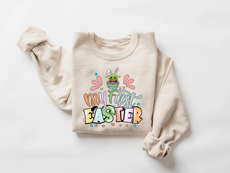 Puede incluir: Sudadera beige con el texto "my first EASTER" en letras de colores. El gr&aacute;fico presenta un personaje de dibujos animados con orejas de conejo en un huevo de Pascua. Acentos rosas y azules rodean el texto.